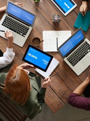 Group of marketers collaborating at a wooden table with laptops and a tablet displaying performance metrics, emphasizing strategic digital marketing and data-driven results.