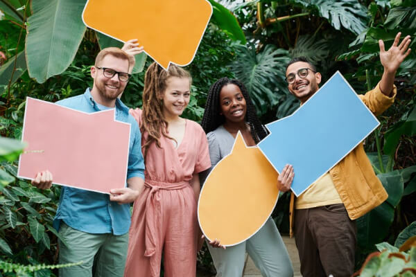 Group of diverse individuals holding colorful speech bubble cutouts in a lush green setting, symbolizing communication and collaboration in digital marketing.