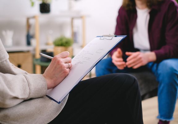Therapist taking notes on clipboard while engaging with a client in a comfortable counseling setting, emphasizing mental health support and connection.
