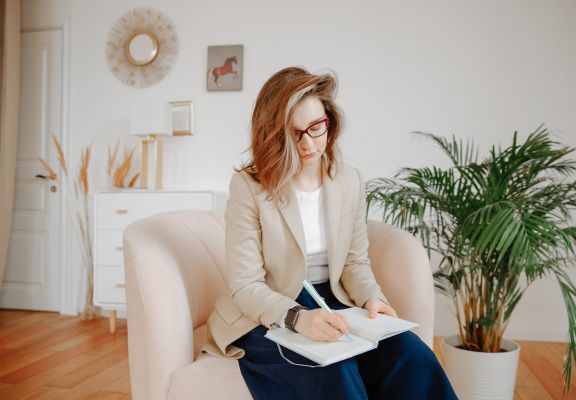 Woman in a light blazer sitting on a plush chair, writing in a notebook, surrounded by indoor plants and a modern, calming interior, reflecting a therapeutic and supportive environment for mental health practices.