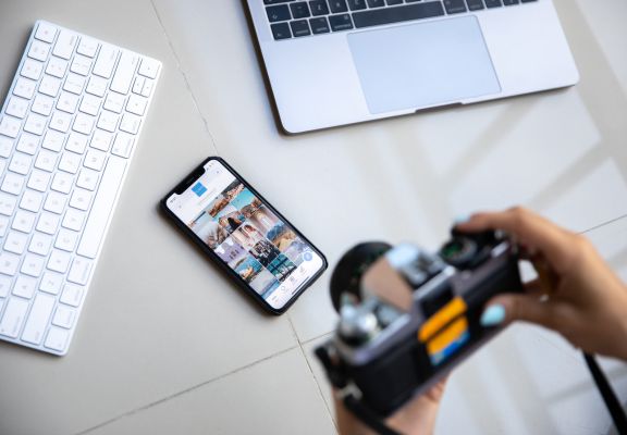 Person holding camera above smartphone displaying photo gallery, with laptop and keyboard in the background, illustrating digital marketing and content creation.