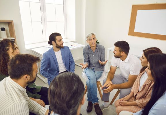Group coaching session with diverse participants engaged in discussion, led by a facilitator in a casual setting, emphasizing collaboration and personal growth.