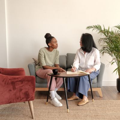 Two women engaged in a therapy session, one taking notes while the other speaks, in a cozy, welcoming setting with a plant and modern furnishings, reflecting a supportive environment for mental health discussions.