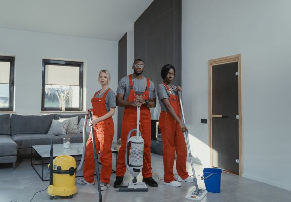 Three diverse cleaning professionals in red overalls standing confidently in a modern living room, equipped with cleaning tools including a vacuum cleaner, mop, and bucket, showcasing home service expertise for local clientele.