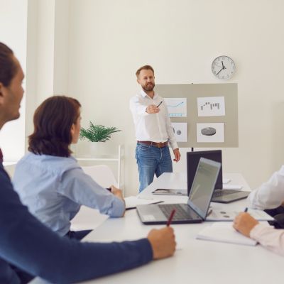 Business meeting with a male presenter gesturing towards a presentation board, surrounded by engaged participants at a conference table with laptops, emphasizing coaching strategies and client engagement.