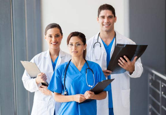 Three healthcare professionals in scrubs and lab coats, smiling and holding clipboards, representing trust and accessibility in the medical field.