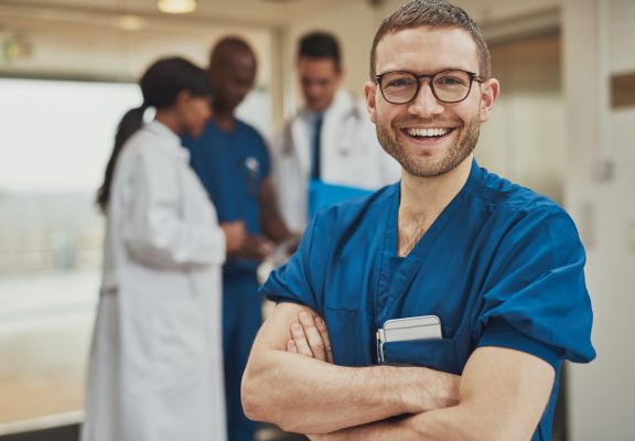 Smiling healthcare professional in blue scrubs with crossed arms, surrounded by diverse medical staff in a clinical setting, emphasizing teamwork and patient-focused care in healthcare marketing.