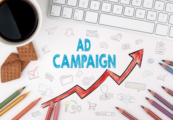 Coffee cup, keyboard, and colorful pencils surrounding a graphic with "AD CAMPAIGN" and an upward trend arrow, symbolizing effective advertising strategies and digital marketing growth.