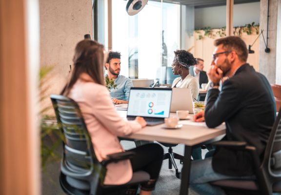 Group of diverse professionals collaborating in a modern office setting, discussing digital marketing strategies with a laptop displaying analytics and charts on the table, emphasizing teamwork for local SEO and business growth.