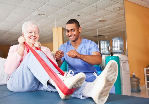 Elderly woman engaging in physical therapy with a male therapist, using a resistance band in a supportive rehabilitation environment.