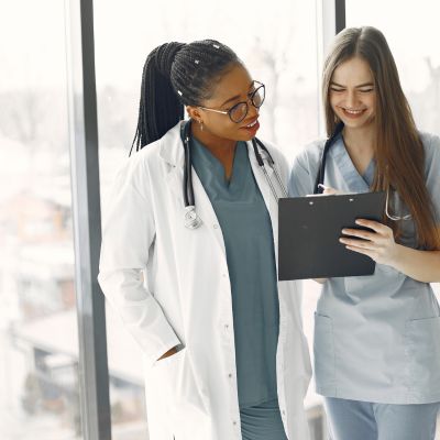 Two healthcare professionals discussing patient information, one in a white lab coat with a stethoscope and the other in scrubs, holding a clipboard, in a bright clinical setting.